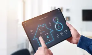 Une main utilise une tablette pour contrôler une application domotique gratuite. L'interface affiche la température, la lumière et d'autres fonctions de la maison.