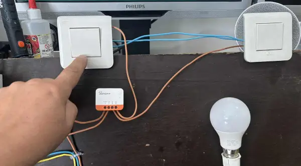 Installation d'un interrupteur zigbee va et vient. Un doigt actionne un interrupteur connecté à un module Sonoff, pilotant une ampoule.