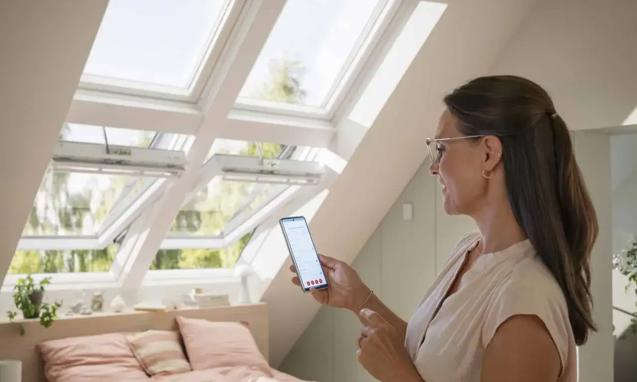 Une femme utilise son smartphone pour piloter des fenêtres de toit Velux, apportant confort et modernité à sa chambre.