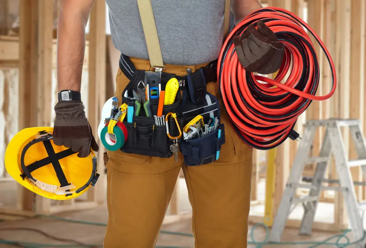 Un &eacute;lectricien porte une ceinture &agrave; outils bien garnie, un casque jaune et tient un long c&acirc;ble rouge et noir.