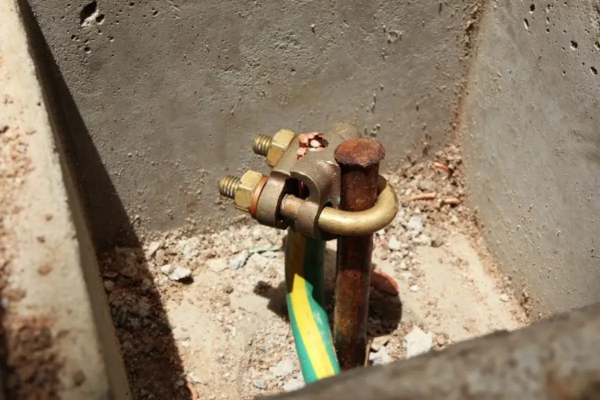 Connexion de mise &agrave; la terre obligatoire. Un collier en laiton relie un fil vert/jaune &agrave; une tige de terre rouill&eacute;e, fix&eacute;e dans du b&eacute;ton.