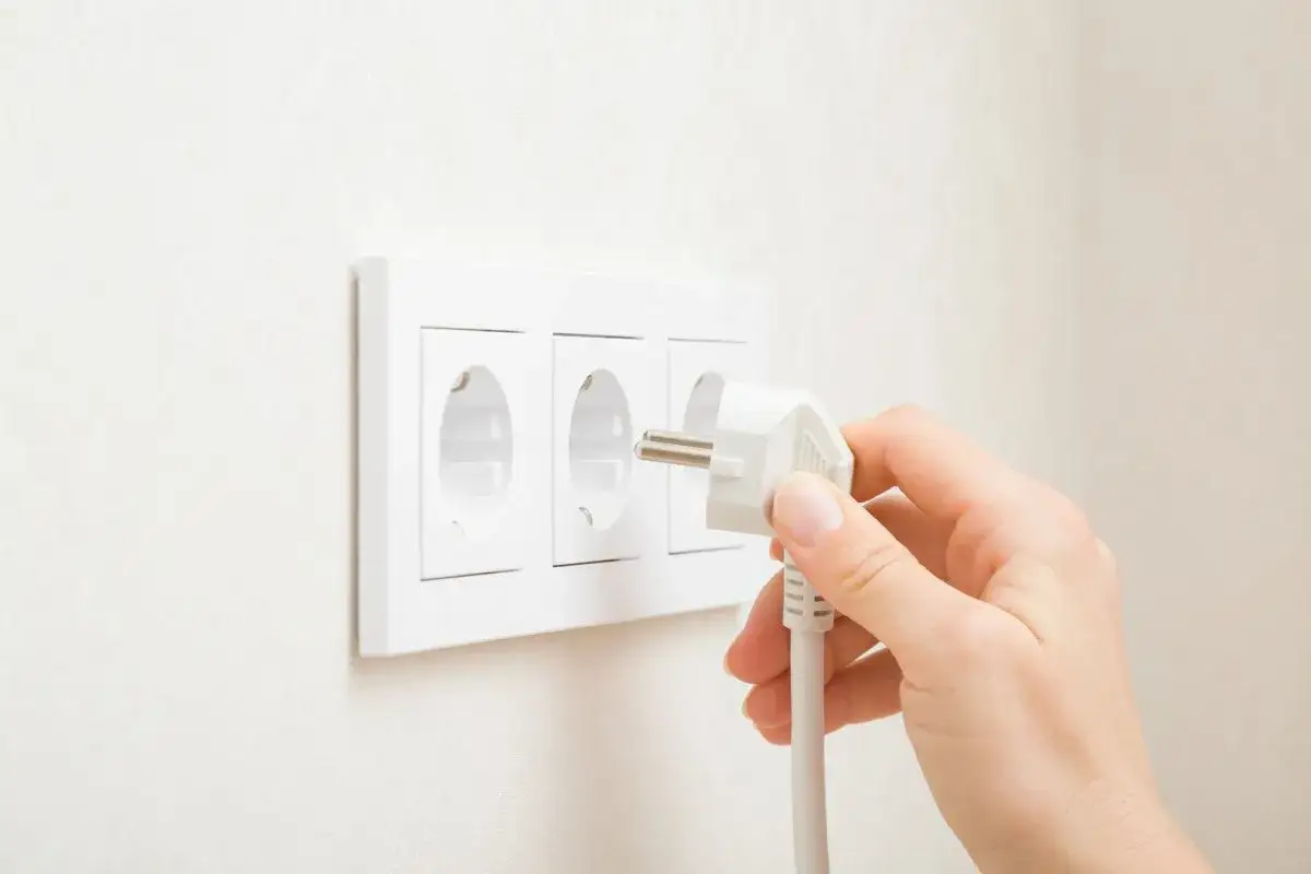 Main d'une personne ins&eacute;rant une prise dans une prise &eacute;lectrique murale triple. La prise est blanche, avec des broches m&eacute;talliques visibles. La prise murale est &eacute;galement blanche et fix&eacute;e sur un mur textur&eacute;. La sc&egrave;ne &eacute;voque la connexion pour la puissan...